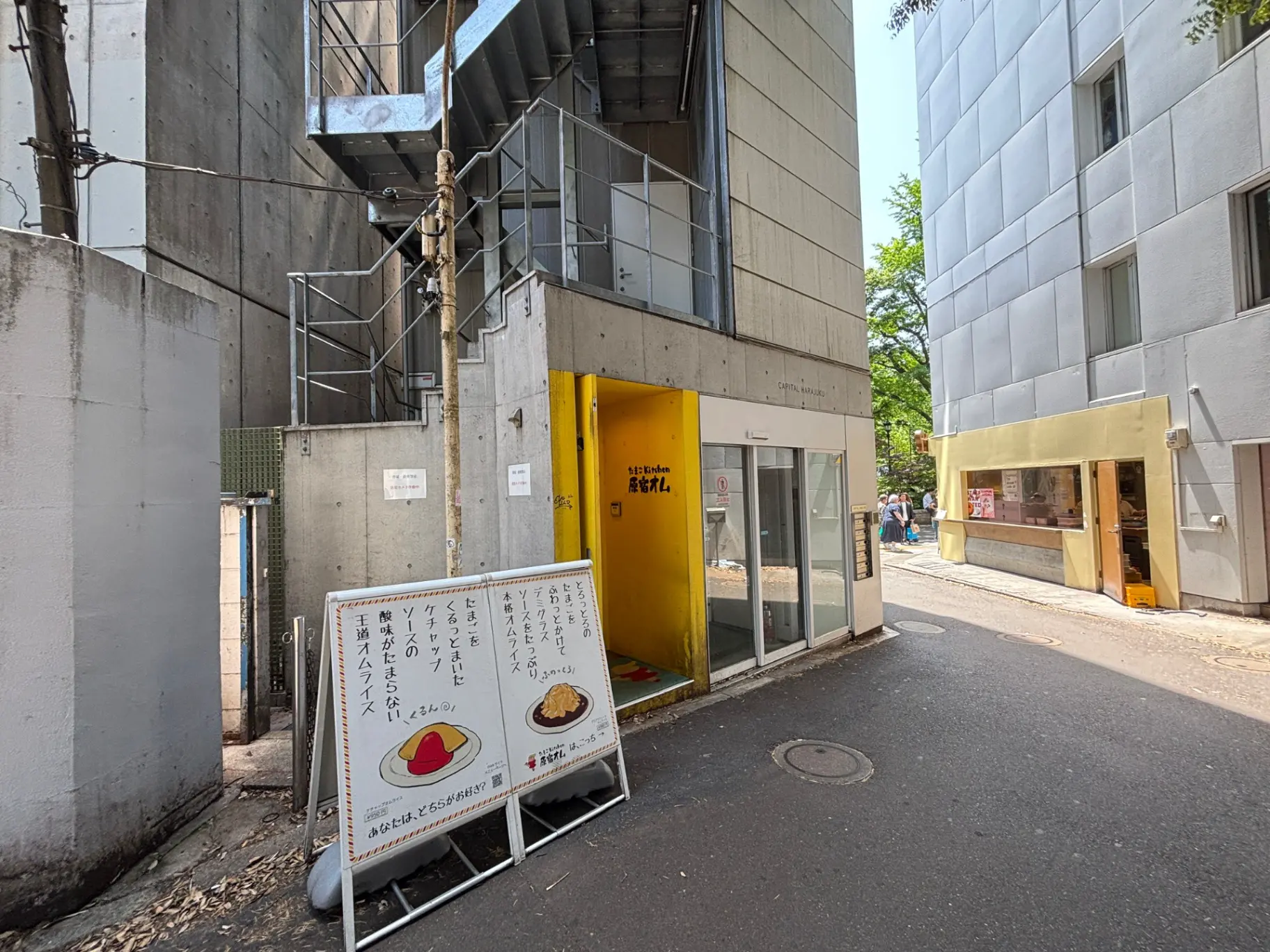 - コンクリート造の建物と明るい黄色の入口、看板には美味しそうな料理のイラストが描かれた飲食店の外観