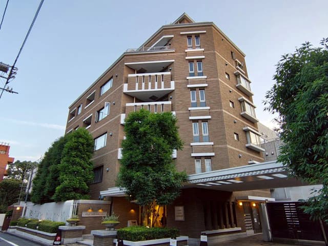 - モダンな外観の高層マンション、緑豊かな植栽とエントランス、晴れた青空の下での夕方の風景