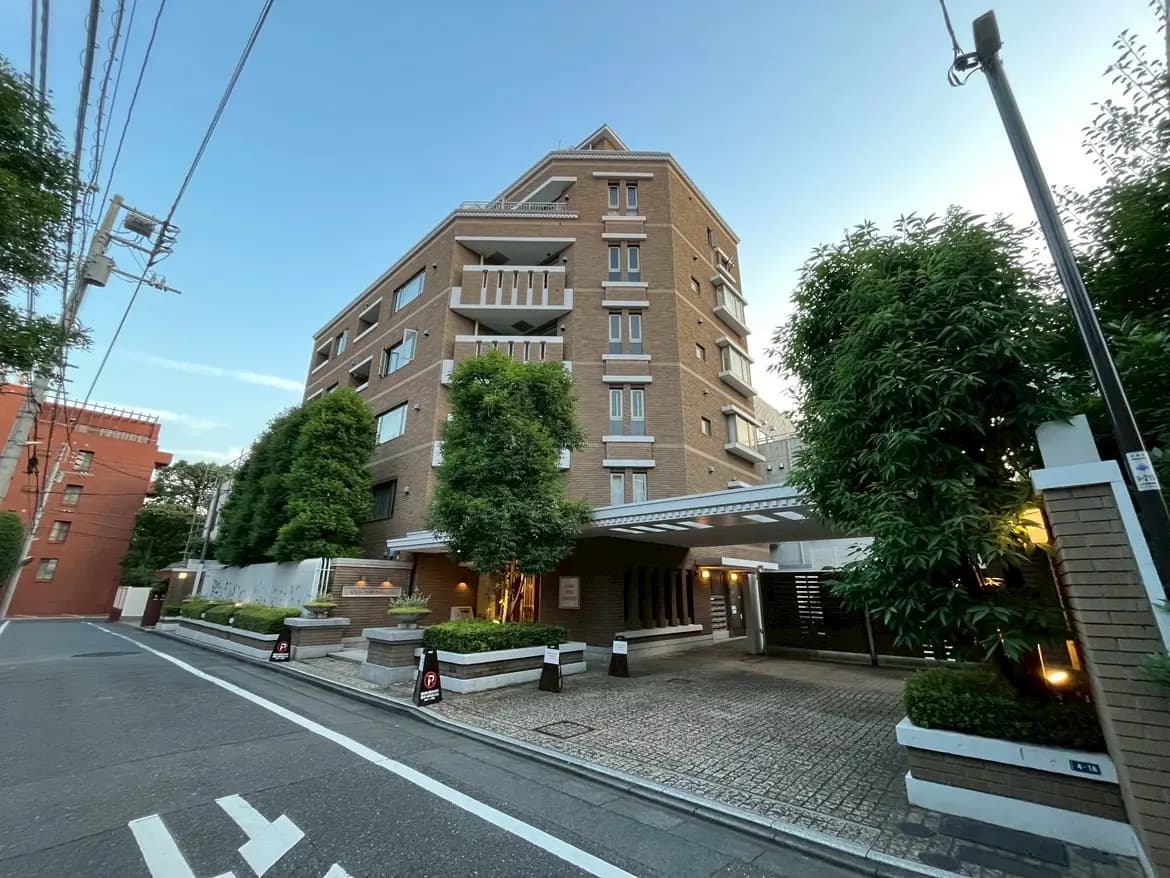 モダンなデザインのマンション、緑豊かな植栽と照明、周囲の街並みが映る外観