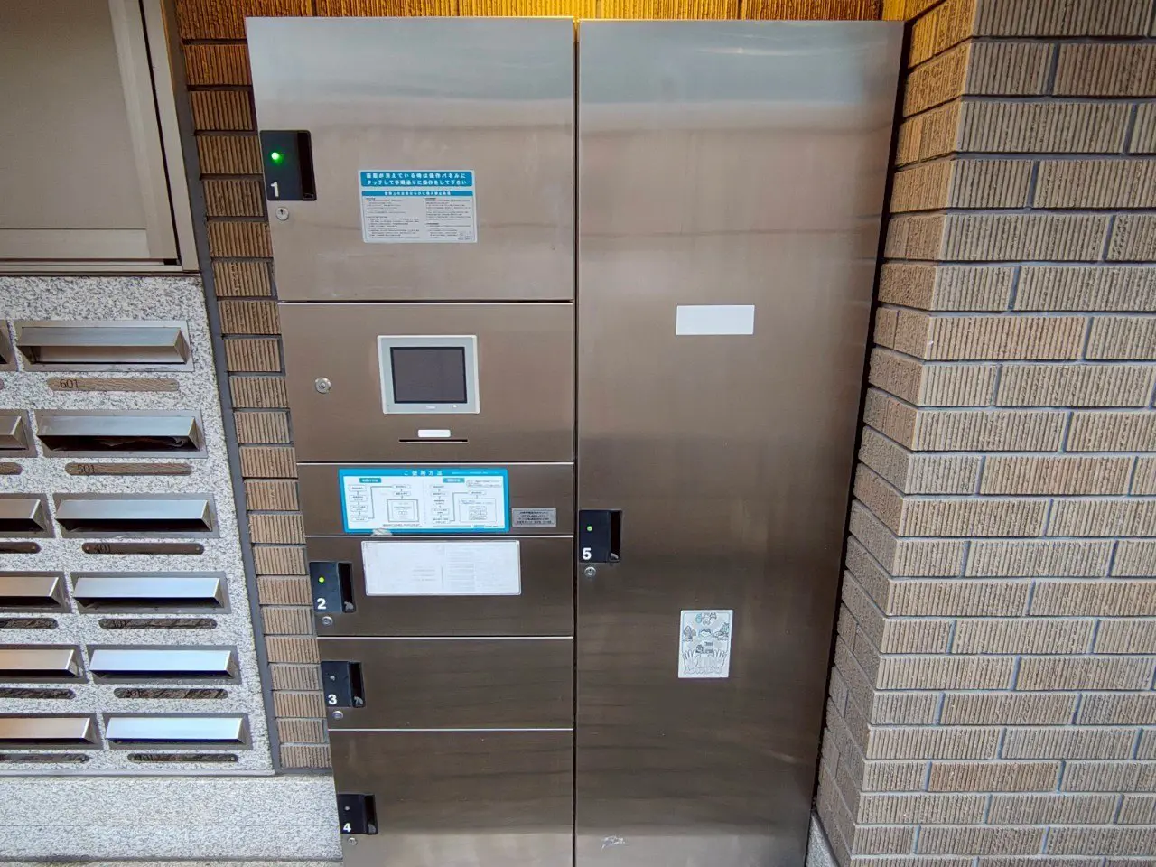 - 銀色の金属製宅配ボックス、上部にデジタルディスプレイと操作パネル、横には郵便受けが並んでいるエントランスの一部