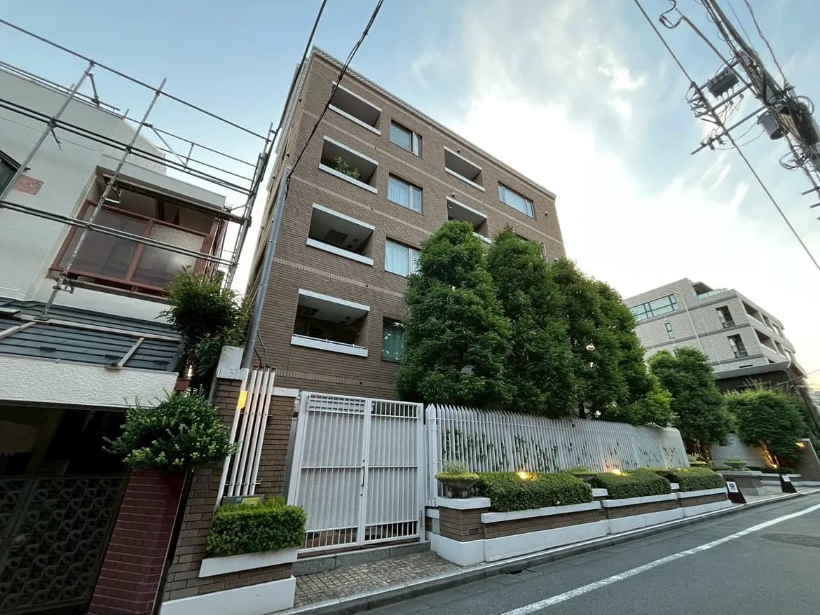 - モダンな外観のアパートメントビル、緑豊かな植栽と白いフェンス、周囲の街並みが映える風景