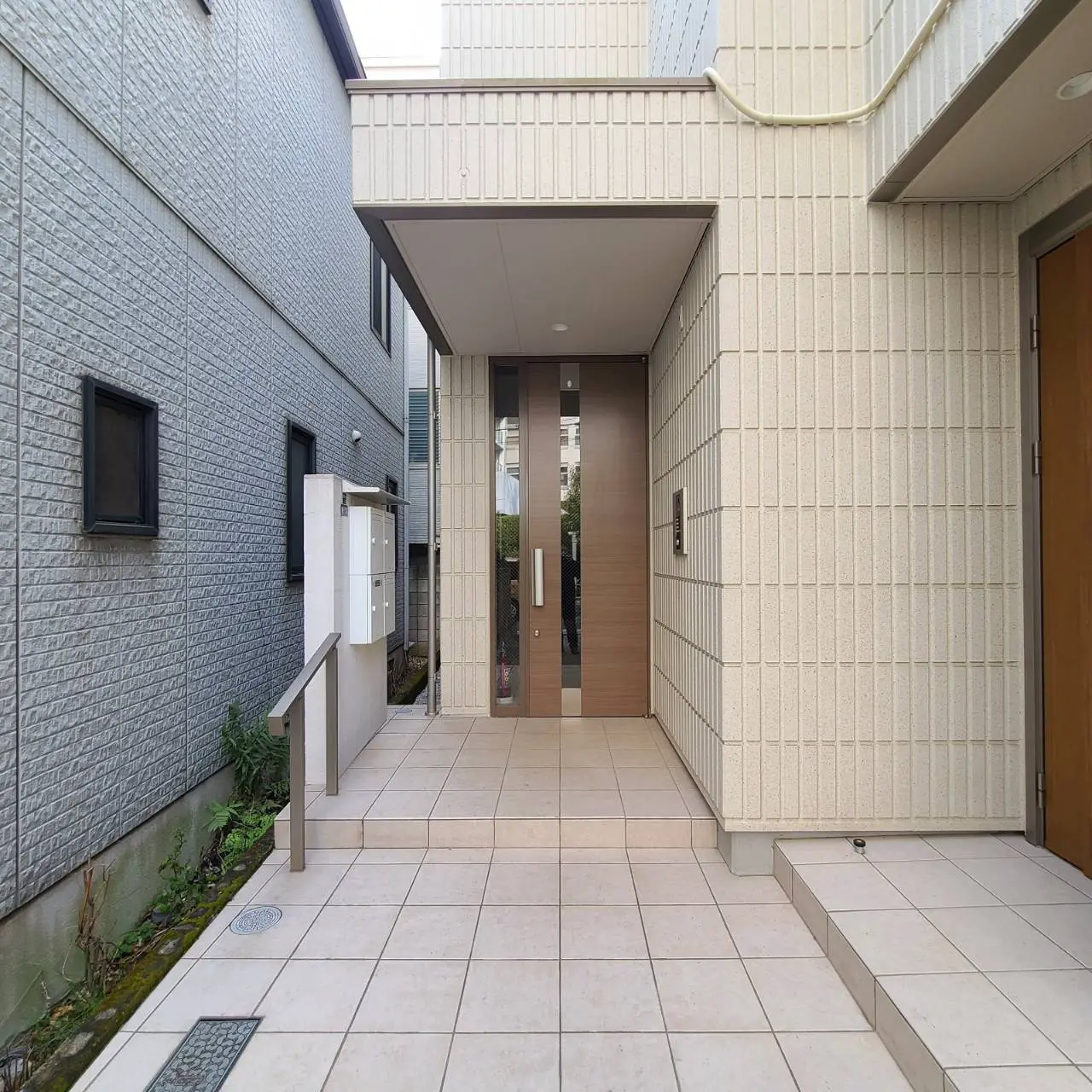 現代的な建築スタイルのエントランス、明るい色のタイルが敷かれたアプローチ、両側に壁がある住宅の外観