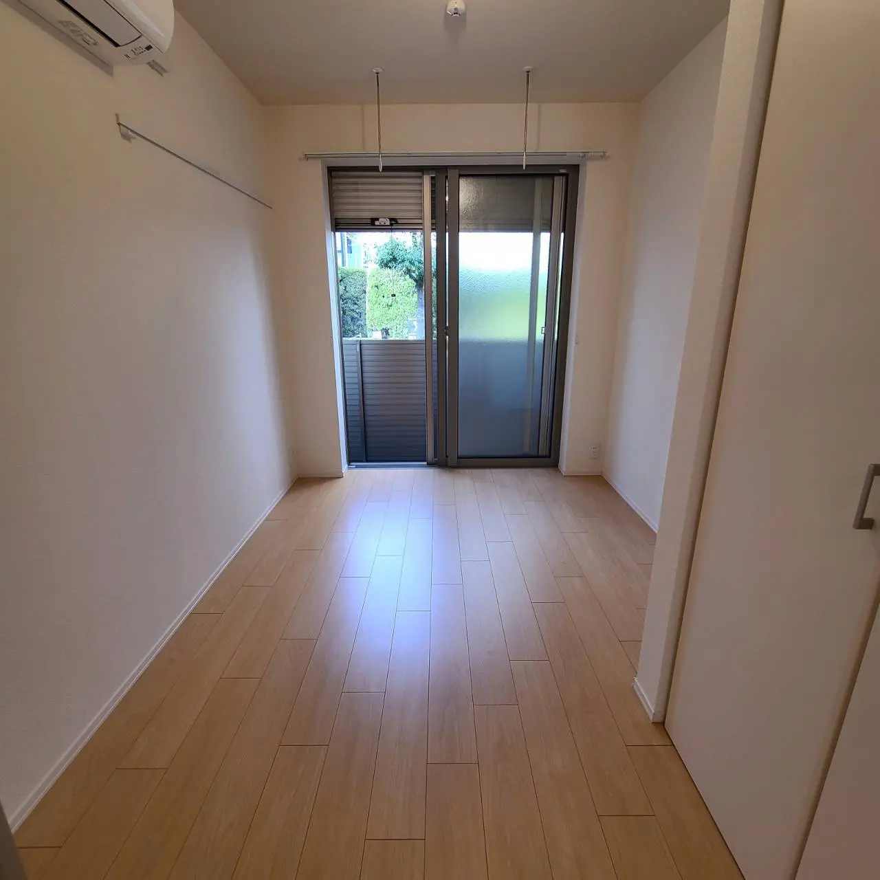 明るく清潔な印象の空の部屋、木目調フロアと白壁、バルコニーへの引き戸とエアコン付き