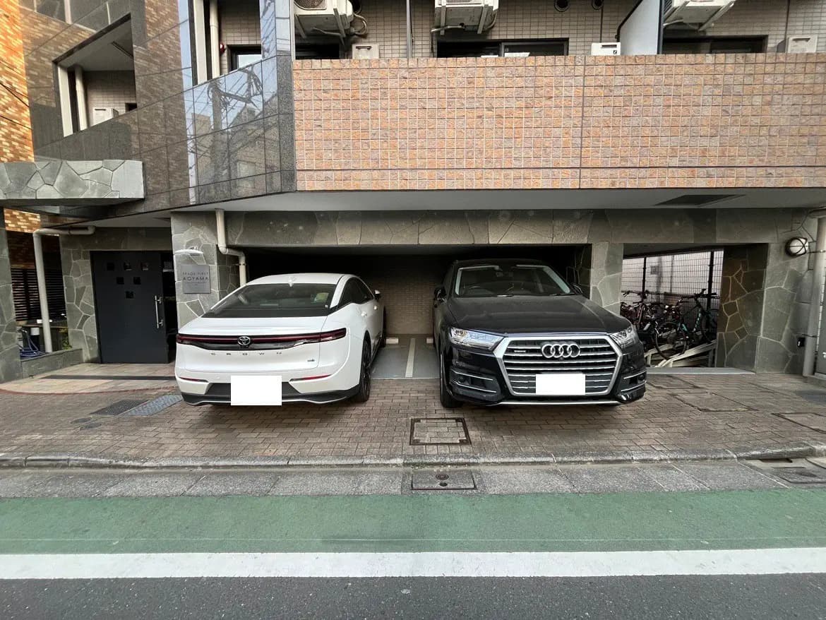 都会的な建物の駐車スペースに停められた白と黒の高級車、背後に自転車置き場が見える外観