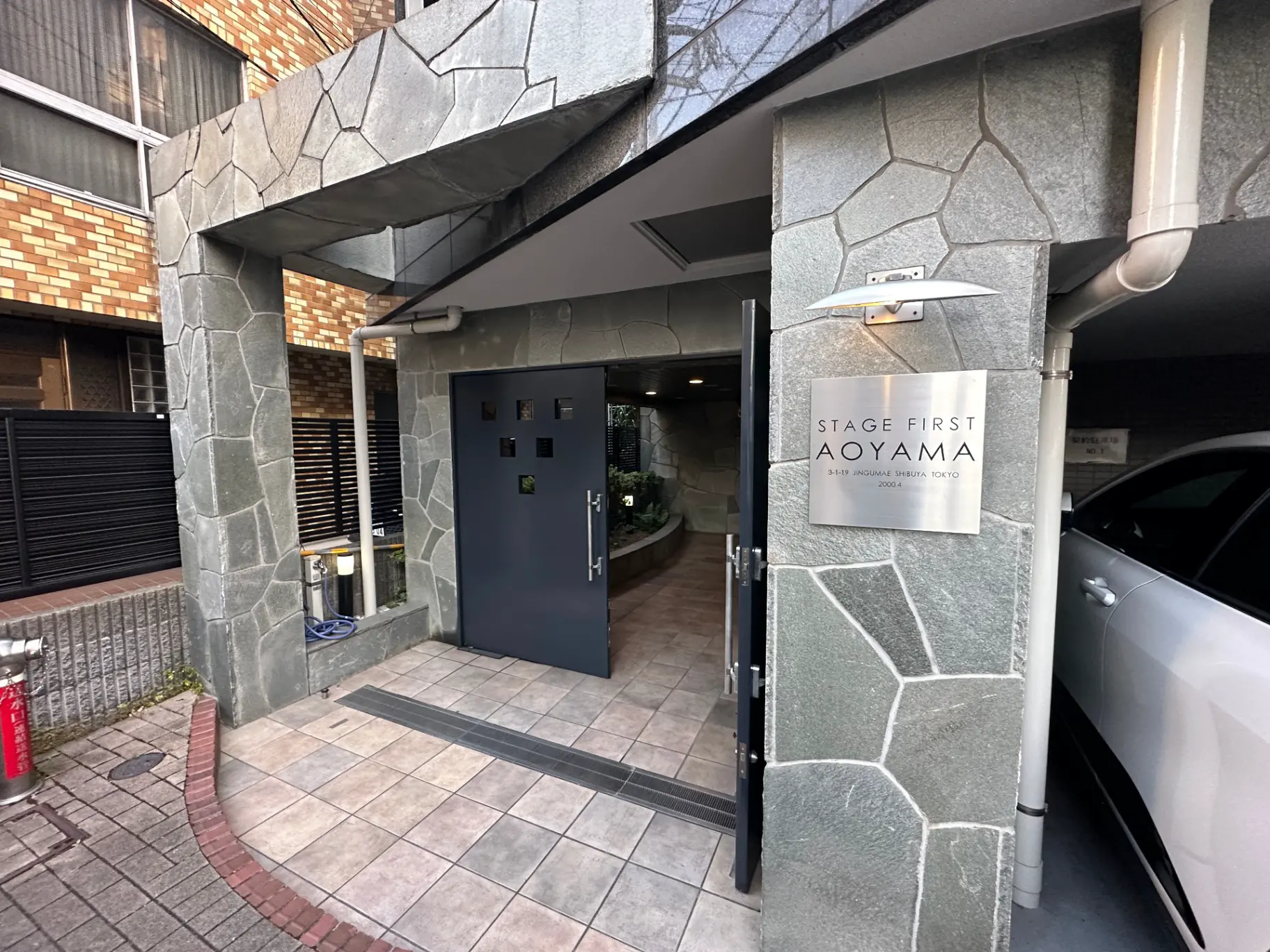 モダンな建築スタイルのエントランス、石の壁面デザイン、STAGE FIRST AOYAMAの看板がある都市のアパート入口
