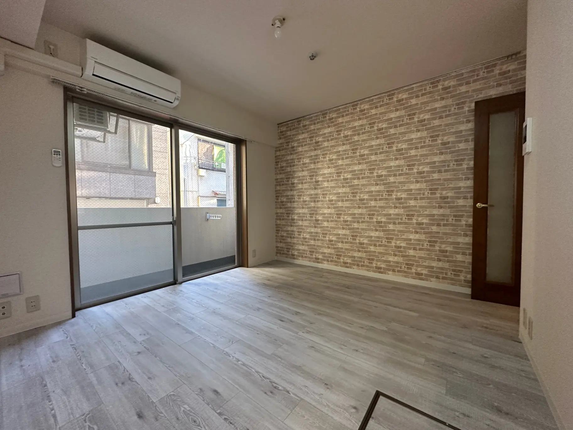 シンプルな内装の空の部屋、レンガ調の壁と木目調フローリング、エアコンと掃き出し窓が付いた明るい室内