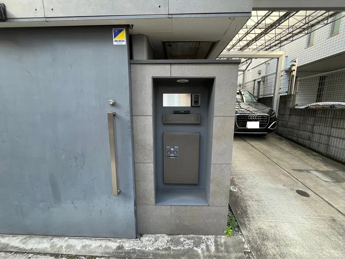 - モダンなデザインの集合住宅のエントランス、専用のインターホンと郵便受けが設置された外壁、隣には駐車スペースに止まっている車の様子