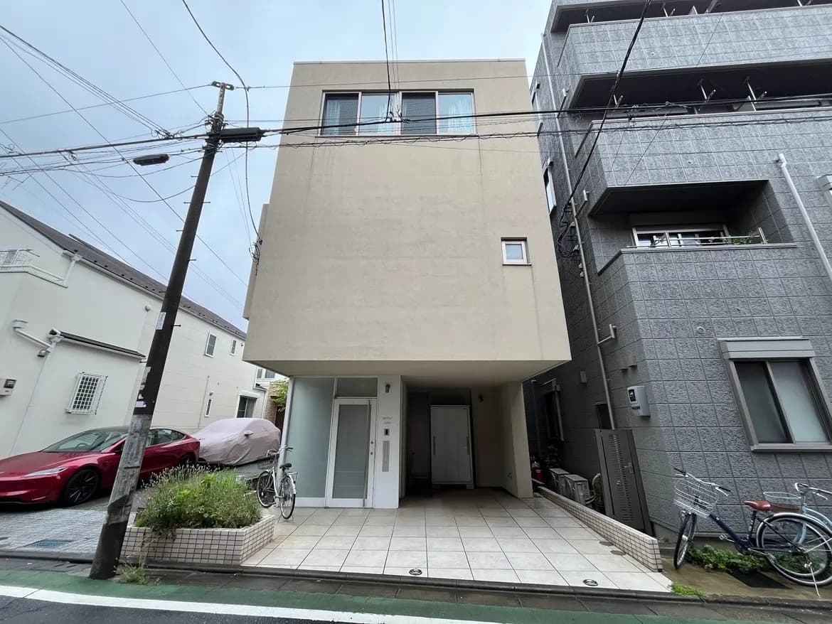 - シンプルでモダンな外観の住宅、隣接する建物と屋外駐車場、電線が交差する都市の風景