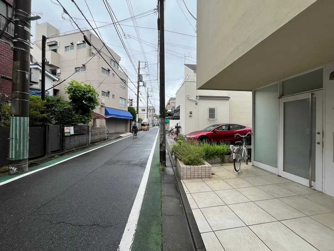 - 雨の日の都市風景、住宅街に面した舗装された道路、駐車中の赤い車と自転車、緑豊かな植栽と近くの建物が見える景観