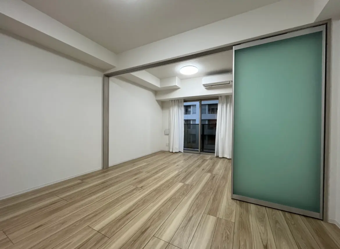 シンプルでモダンなリビングルーム、木目調のフローリング、白い壁、エアコン付き、明るい室内照明、バルコニーへのカーテン付きスライドドア