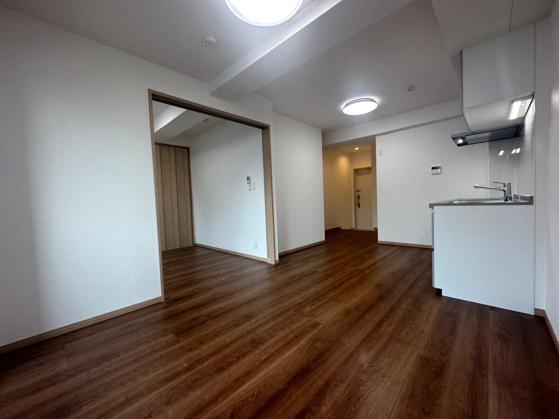 シンプルなデザインの新築マンションの室内、木目調のフローリング、白壁、キッチンスペースとダウンライトが特徴