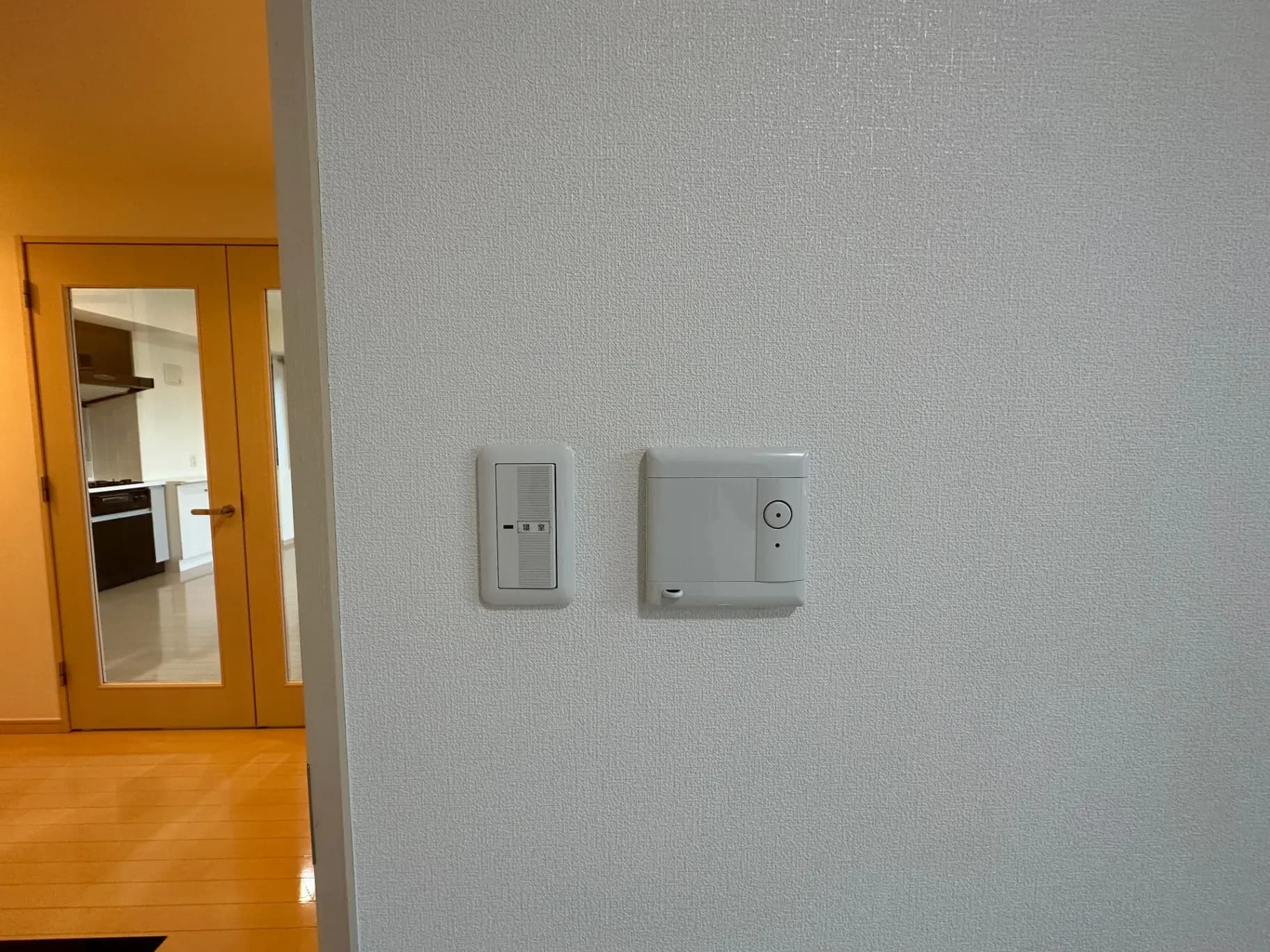 室内の壁に取り付けられたインターフォンとスイッチ、背景に木目調のドアがある明るいエントランス
