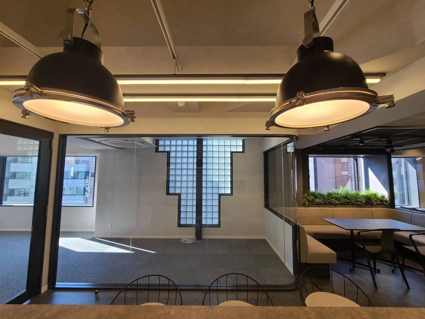 オフィスのミーティングスペースに吊り下げられたモダンなデザインの照明とプラントボックス、テーブルと椅子が配置された快適なコワーキング空間