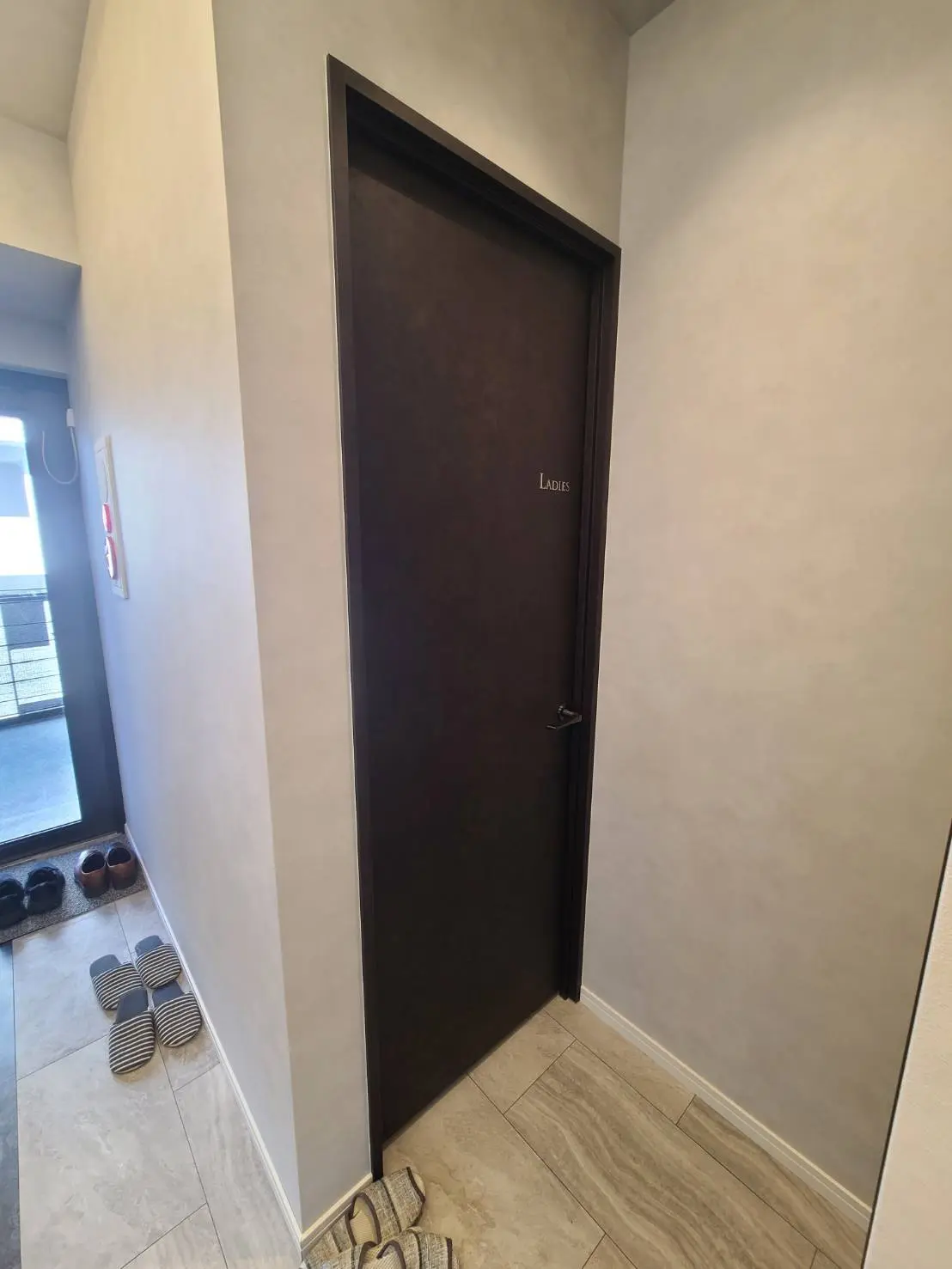 シンプルなデザインの玄関廊下、左側にレディーストイレのドア、床にスリッパと靴