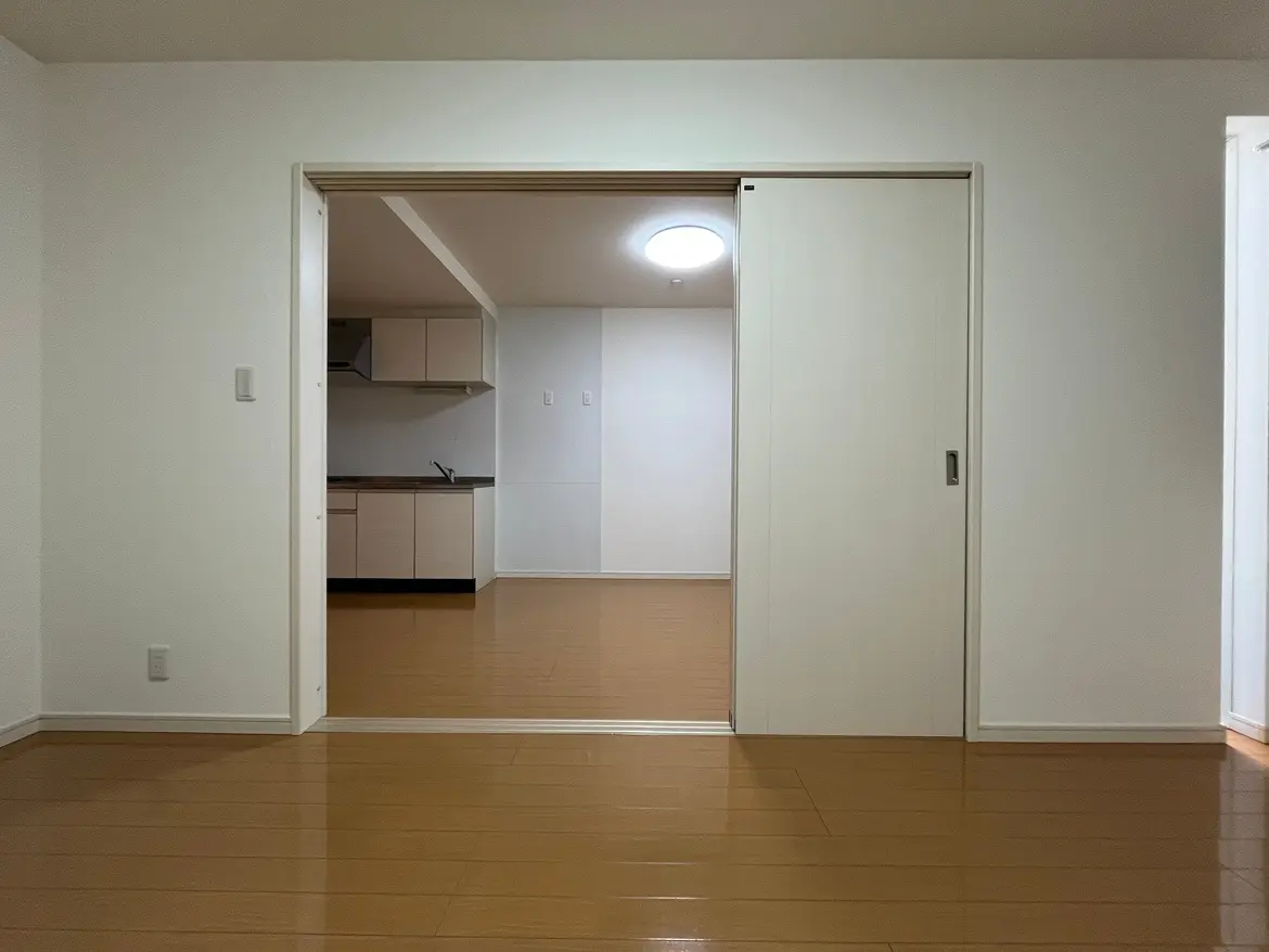 シンプルなデザインの空の部屋、木製フロアと白い壁、キッチンスペースが見える