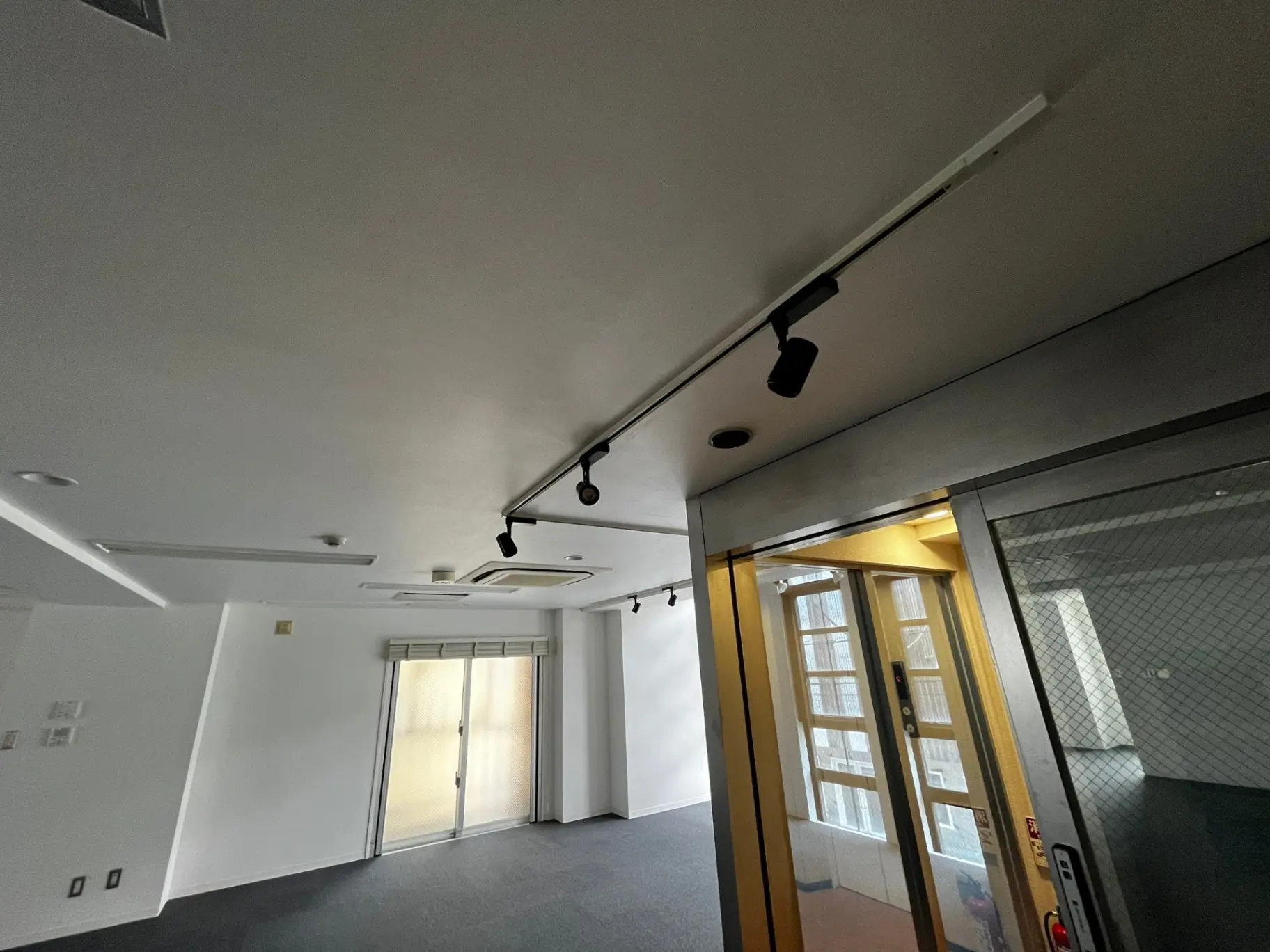 空間が広々としたオフィスの室内、白い壁と天井、黒いスポットライトの照明、ガラス扉のエレベーター入口