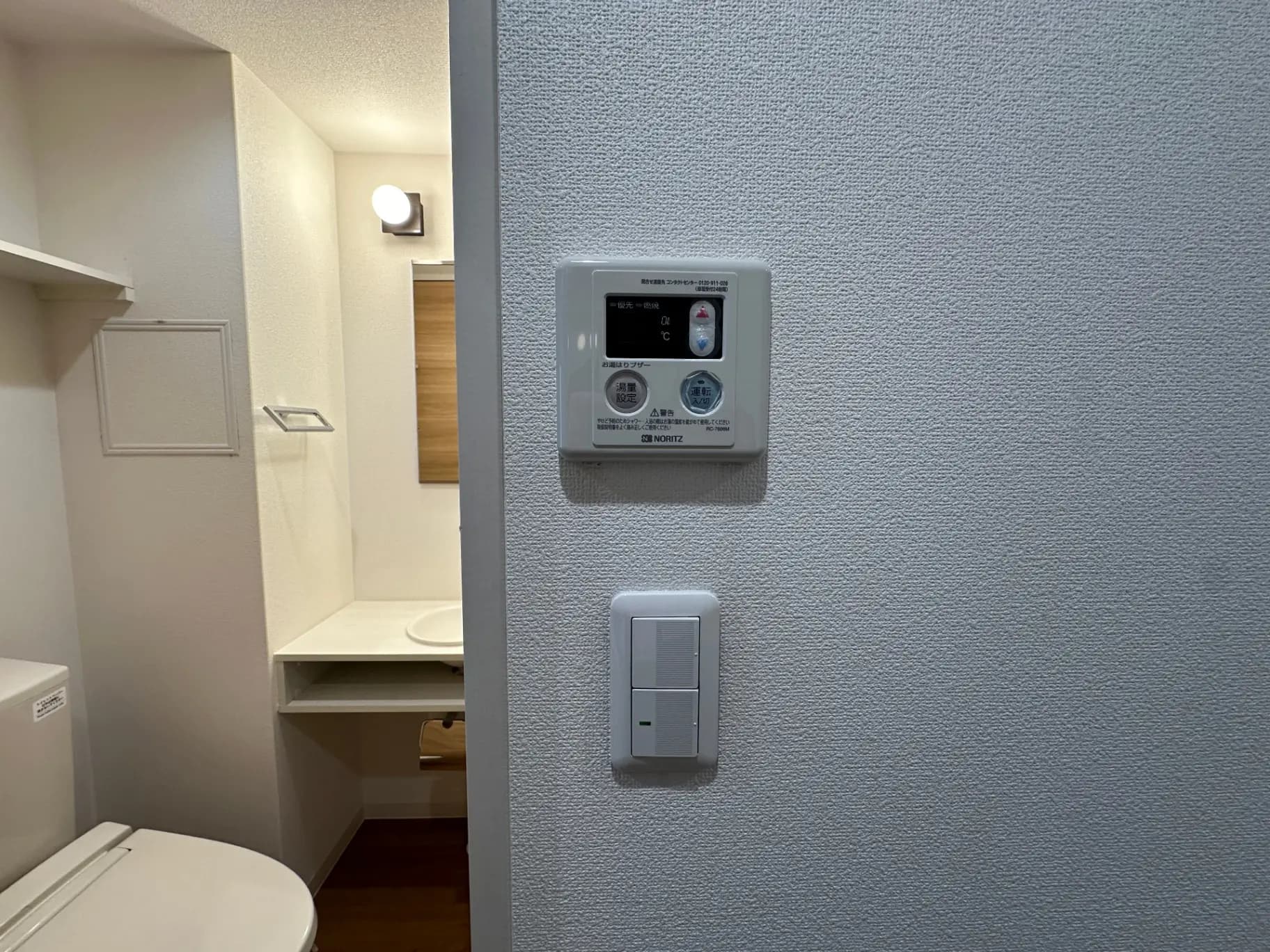 モダンなトイレのインテリア、白い壁にノーリツ製のガス給湯器リモコンとスイッチが取り付けられた壁、木目調の洗面台