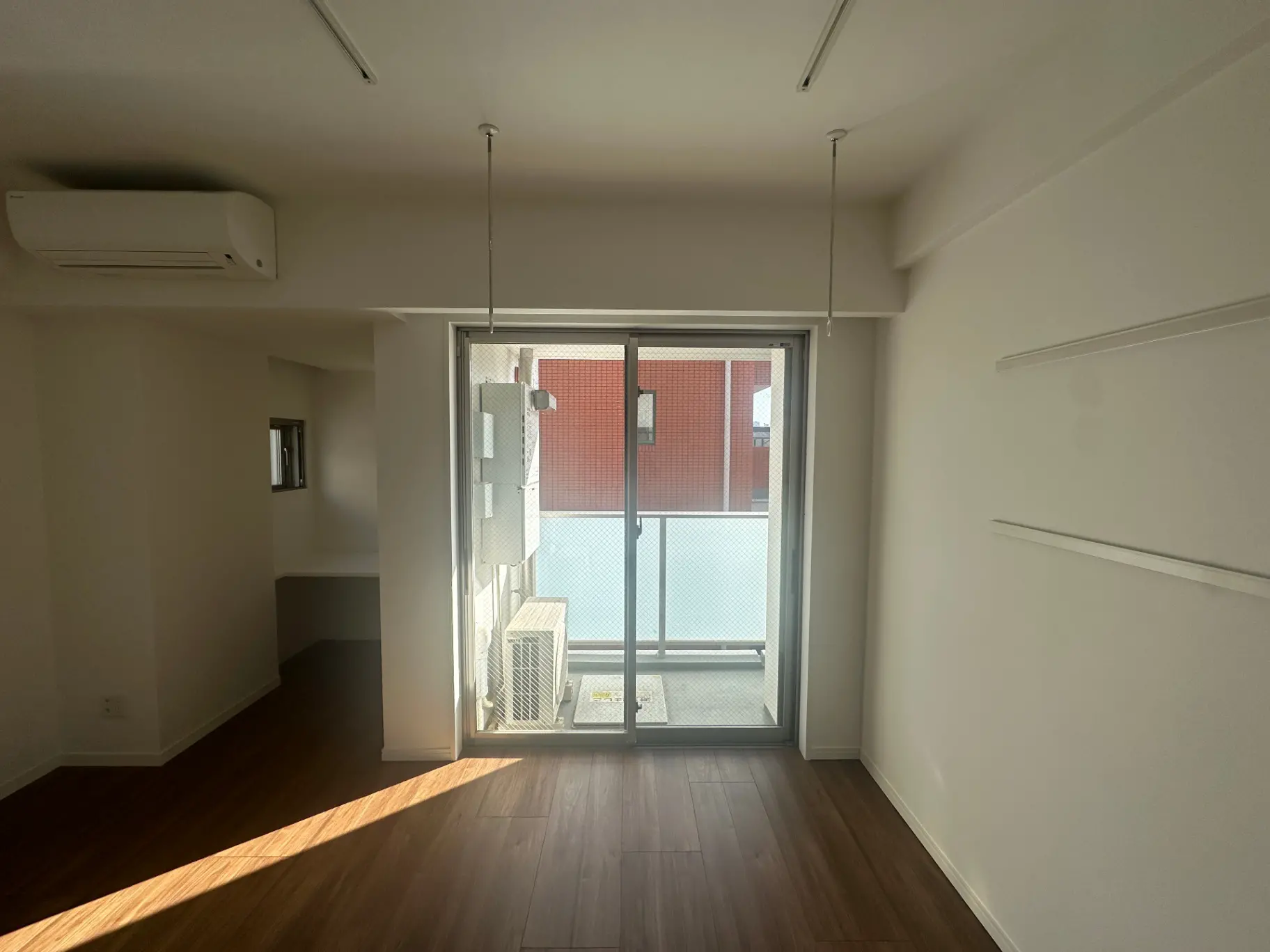 シンプルなデザインの空の部屋、木目調のフローリング、エアコン付き、明るい自然光が差し込むバルコニー付きアパート