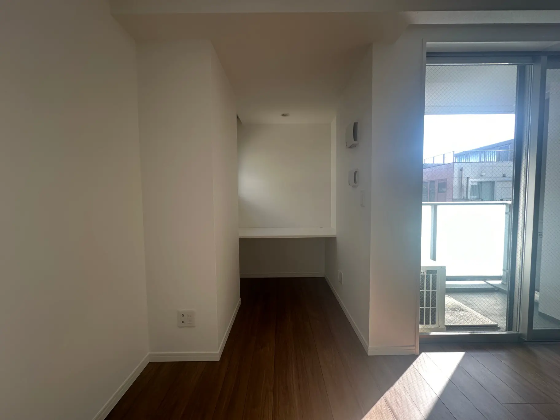 新築マンションの空室、木製フローリングと白い壁、バルコニーへの出入り口付きの明るいリビングルーム