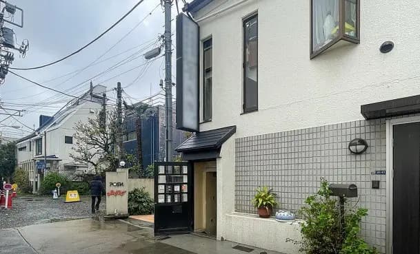 - 雨の日の静かな通りに面した白い外壁の建物、周囲には緑の植物や看板が立ち並ぶ、住宅地の一角の風景