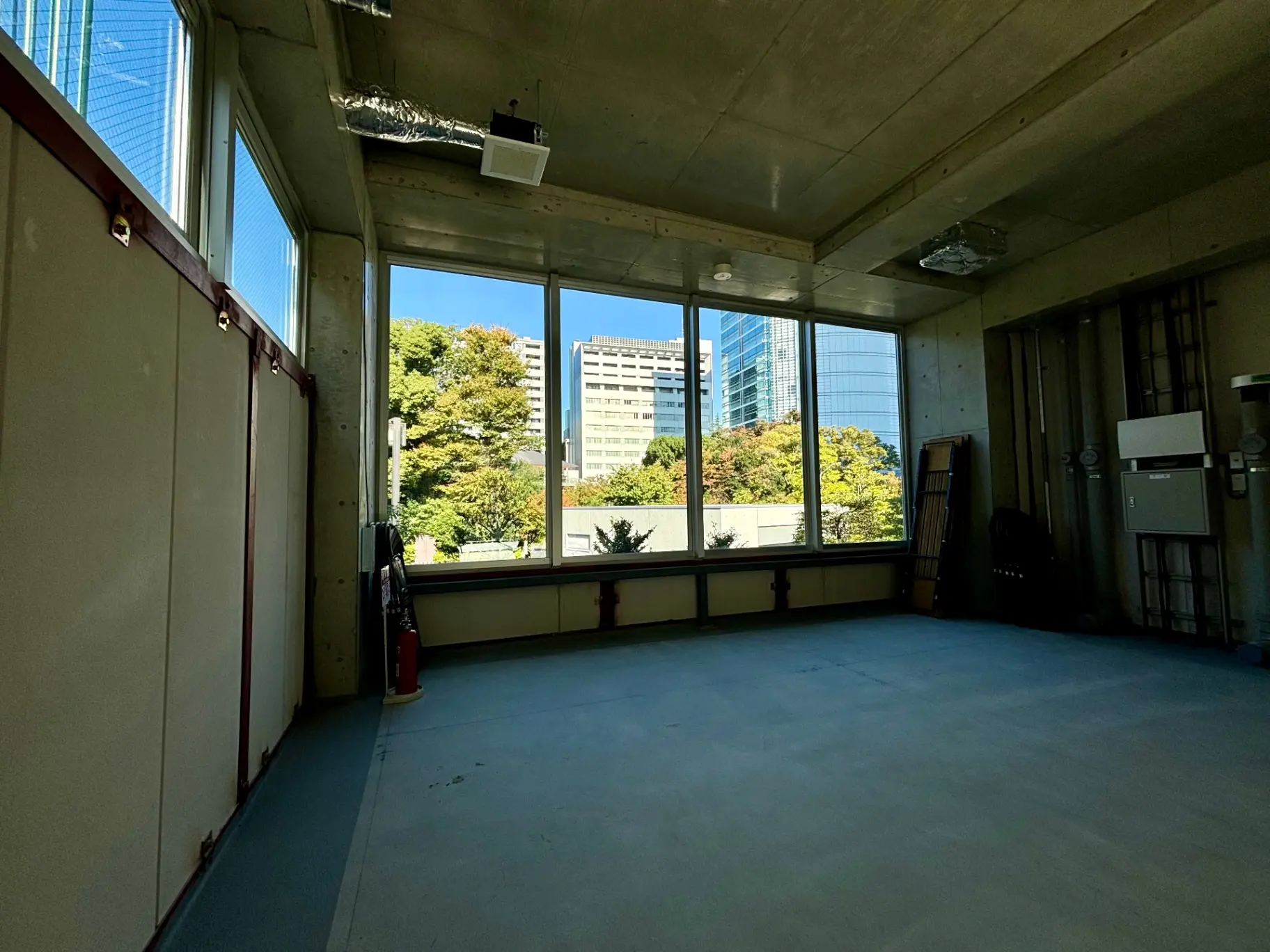- 窓から明るい光が差し込む広々とした空間、コンクリートの天井とシンプルな内装、外には緑豊かな景色と高層ビルが見える室内の一角