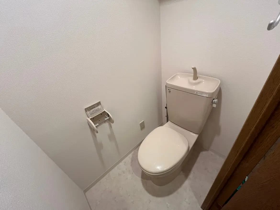 - シンプルで清潔感のあるトイレ、ベージュのトイレ本体と白い壁、手すりが設置された室内の風景