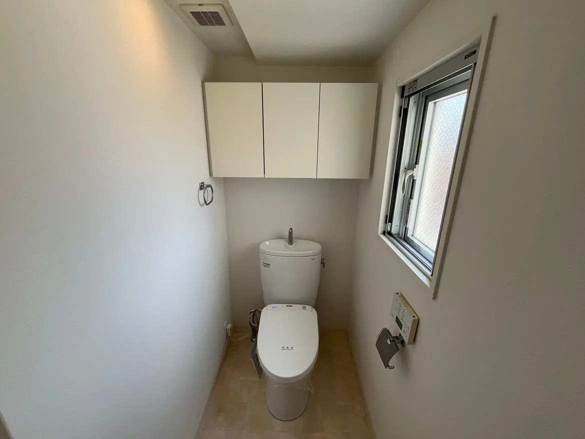 - 明るいトイレのインテリア、白い壁と収納棚、横には窓があり、シンプルなデザインの便器とトイレットペーパー掛けが配置されている空間