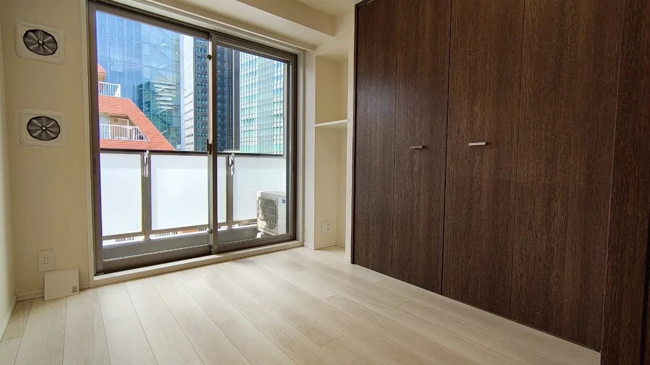 - 明るい室内と大きな窓からの都市景観、シンプルなデザインのウォークインクローゼットと木目調のフローリングが特徴の部屋