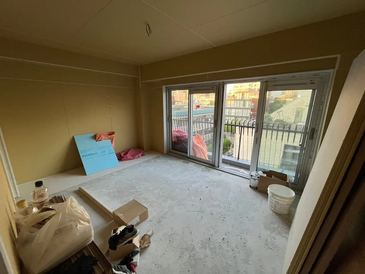 - リフォーム中の部屋、コンクリートの床と未完成の壁、バルコニーに面した大きな窓と雑然とした物品が置かれたインテリア