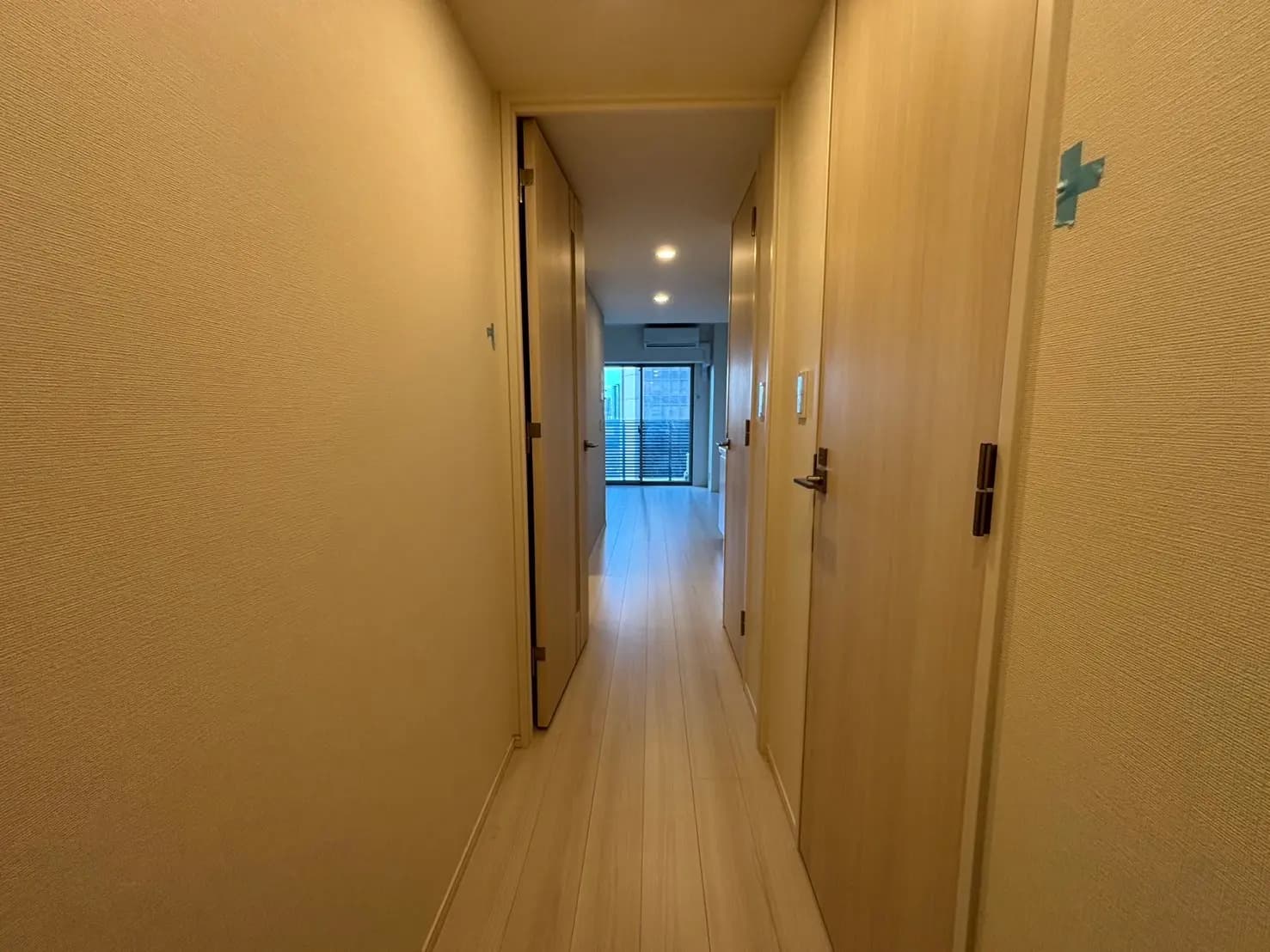 - 明るい色合いの壁と木製のドアが並ぶ住居の廊下、奥には窓からの自然光が差し込むリビングスペースが見えるインテリア