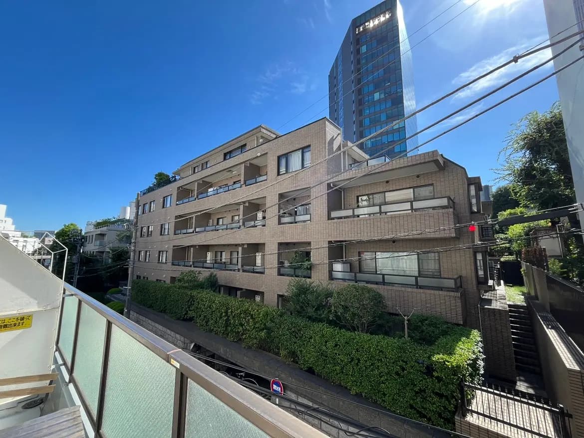 - 都市部に立つモダンなアパートメントビル、青空と緑豊かな植栽が広がる風景とバルコニーが見える外観