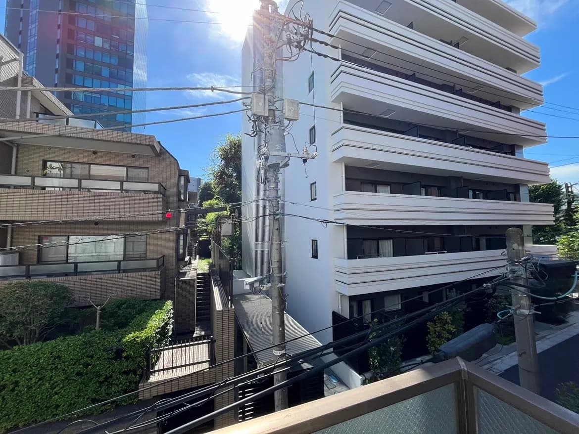- 高層ビルと住宅が立ち並ぶ街並み、青い空の下で明るい日差しが差し込むシーン、電線や緑の植栽が見える風景