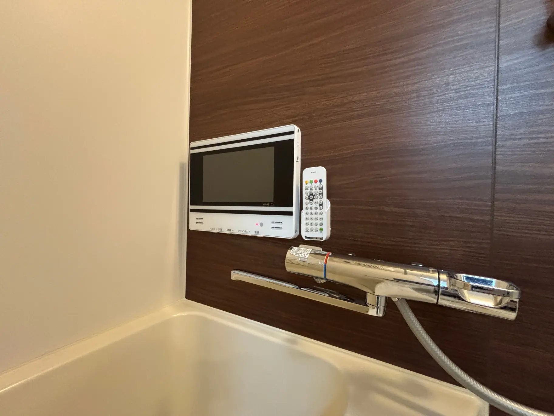 - 現代的なバスルームの壁に取り付けられたテレビモニターとリモコン、ステンレスの蛇口と白い浴槽の一部が写っている画像