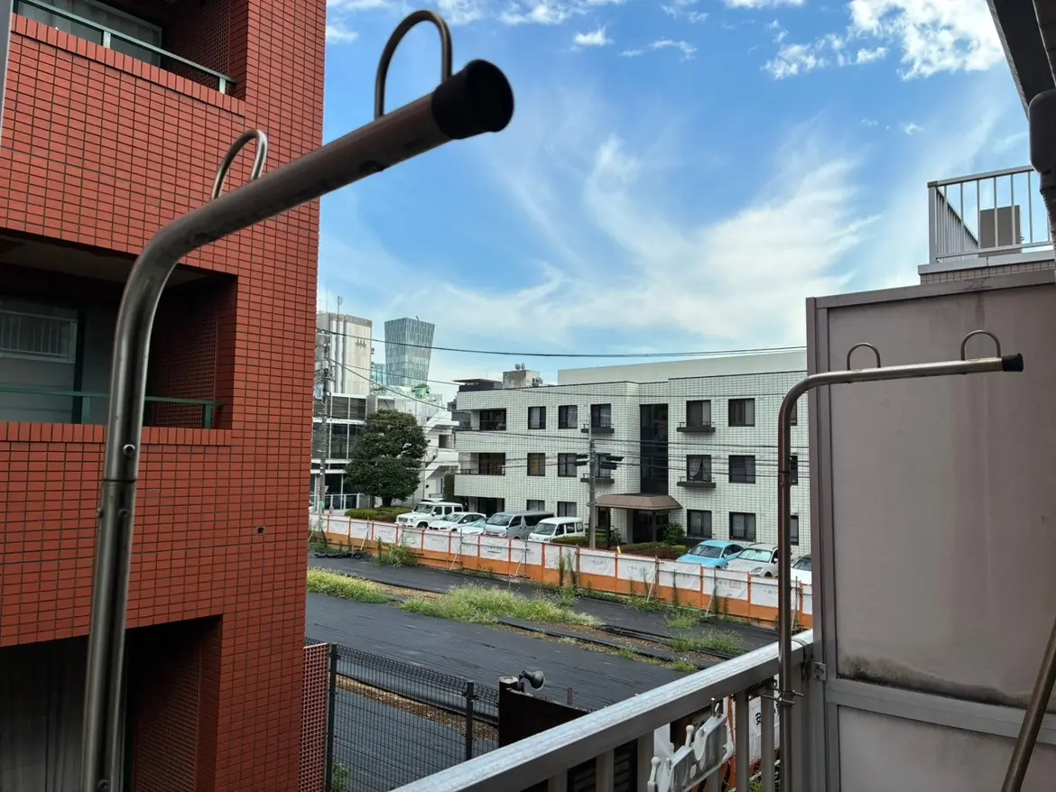 - ベランダから見える都市の風景、隣接する建物と青空に映える雲、洗濯物干し用のステンレス製のポールが写っているシーン