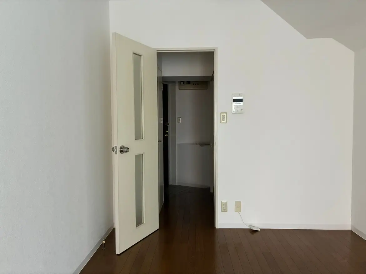 - 開いたドアが見える室内の風景、白い壁と木製のフローリング、隣の部屋に通じる廊下が映るシンプルな空間