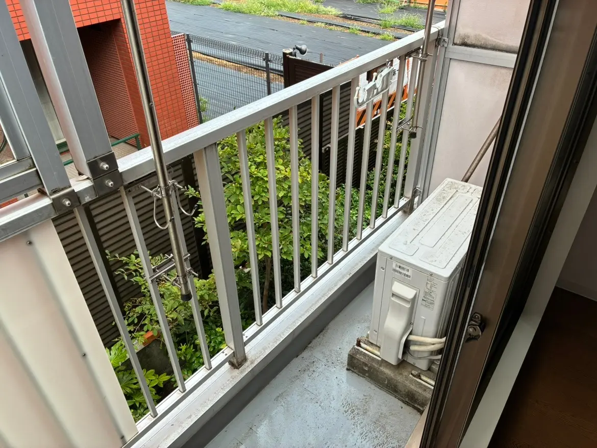 - 鉄製の手すりが付いたバルコニー、背景に緑が広がる植栽とエアコン室外機が置かれている風景