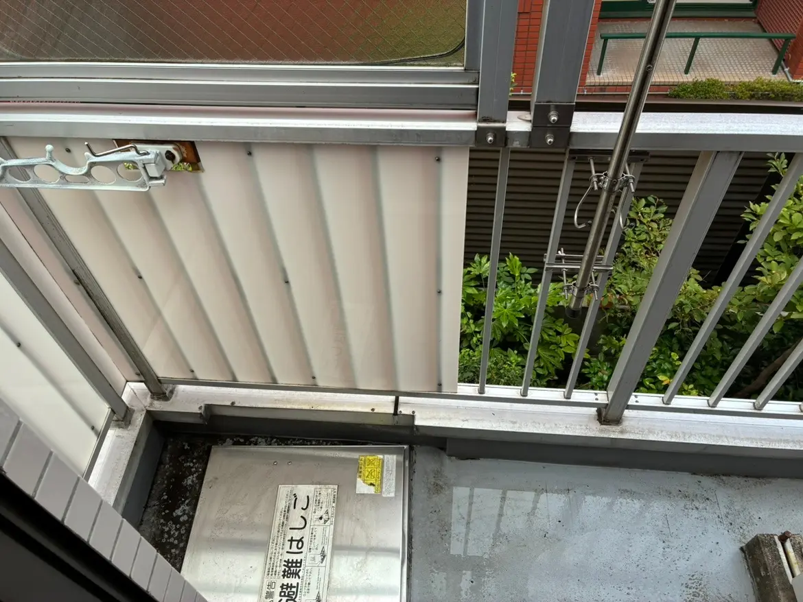 - ベランダの金属製手すりと透明パネルの近up、下部にある掃除が必要な床面と緑の植物が見える景観