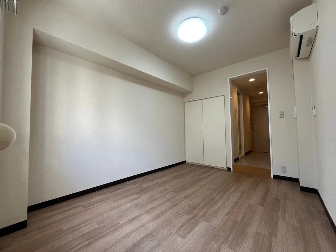 - シンプルで広々とした空間のスタジオタイプの部屋、明るい木目調のフローリングと白い壁、クローゼットとエアコン完備の室内