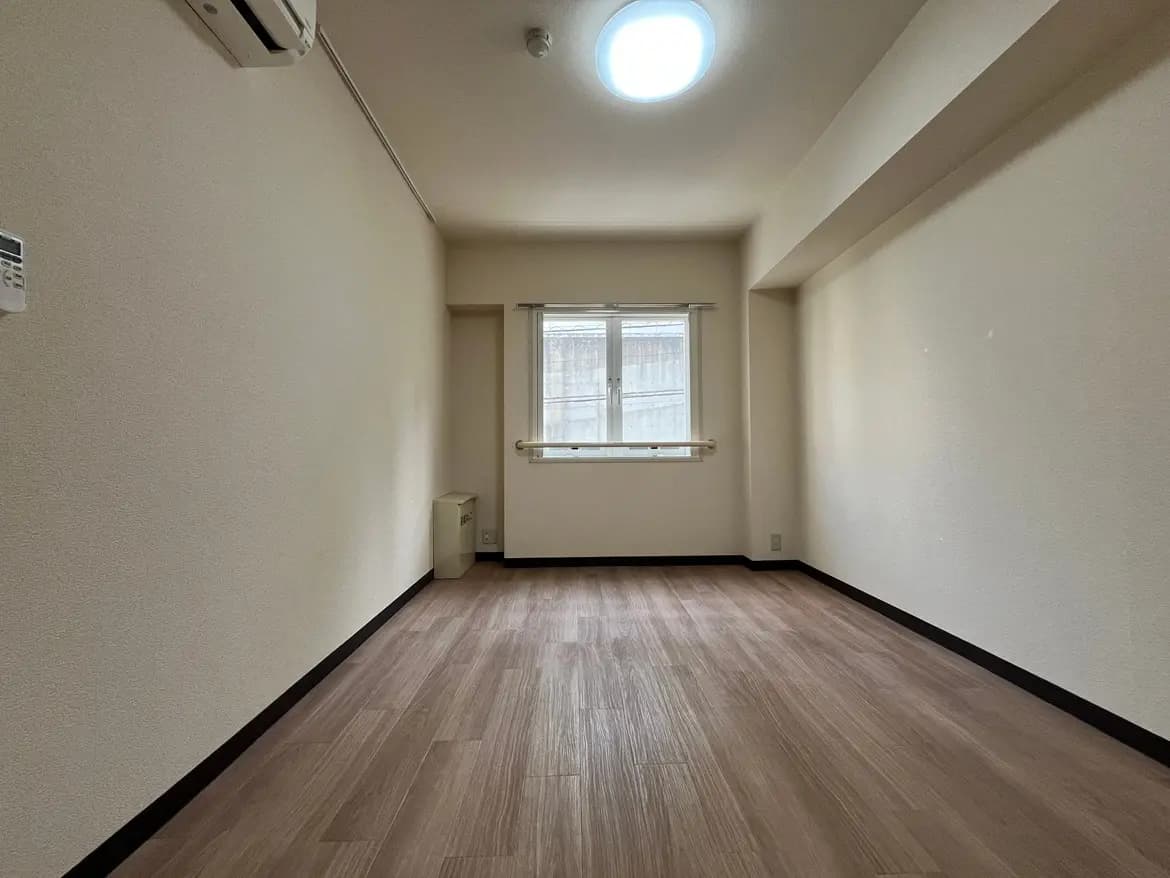 - シンプルで明るい印象の部屋、木目調のフローリング、窓に自然光が差し込むインテリア、エアコンとシンプルな壁面
