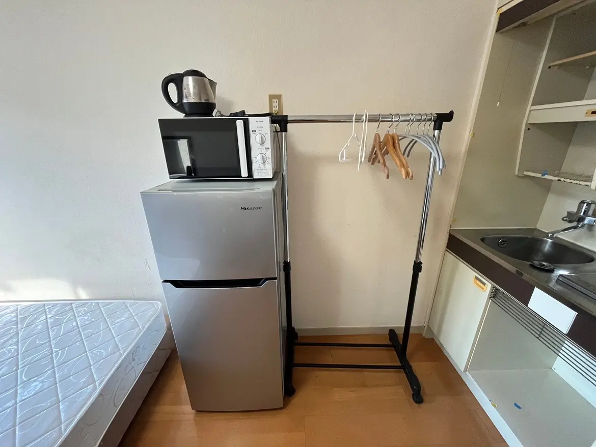 - 薄型冷蔵庫の上に電子レンジとケトルが置かれたキッチンエリア、隣に衣類を掛けるハンガーラックがあるシンプルな部屋の内部