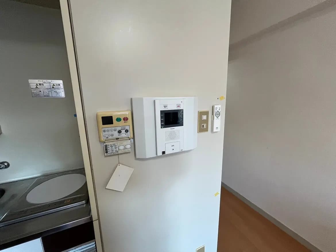 - キッチン横の壁に取り付けられたインターホンとスイッチ類、隣には調理器具が見えるシンプルな室内の一角