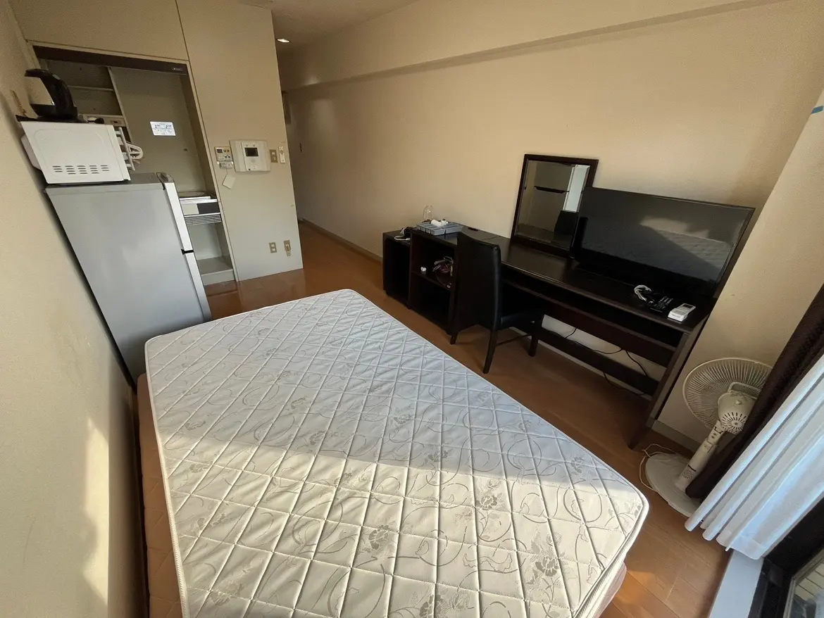 - ナチュラルな木目調のフローリングのシンプルなビジネスホテルの客室、白いベッドマットレス、机とテレビが備え付けられたコンパクトなインテリア