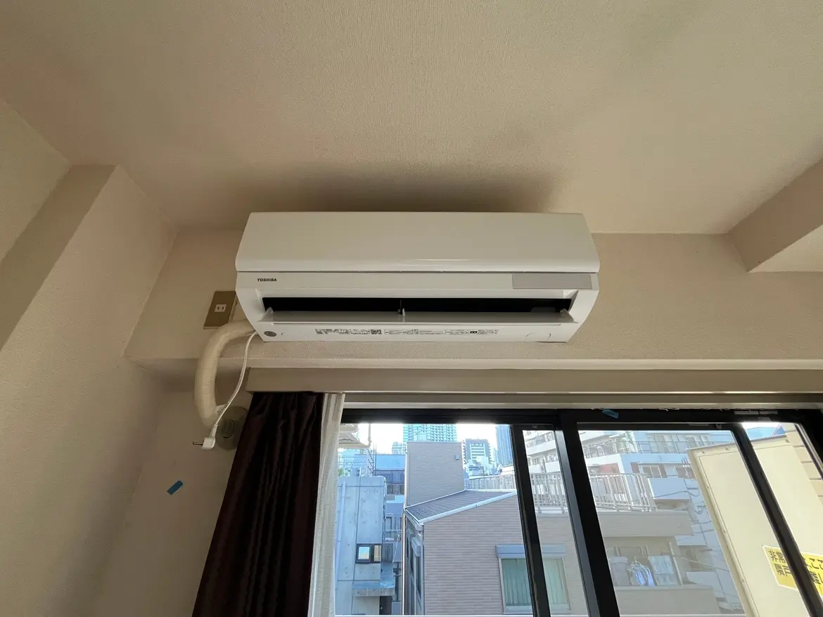 - 部屋に設置された東芝製エアコン、明るい窓とカーテンのあるインテリア、アパートの一室の上部に取り付けられた様子