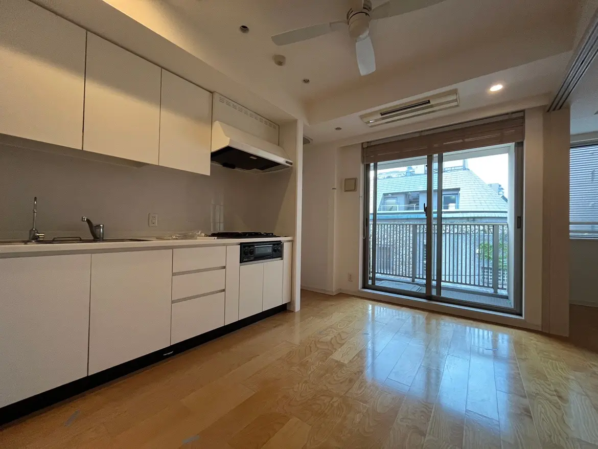 - 白を基調としたシンプルでモダンなキッチン、光が差し込むバルコニー側の大きな窓、木製のフローリングが特徴的な室内のインテリア