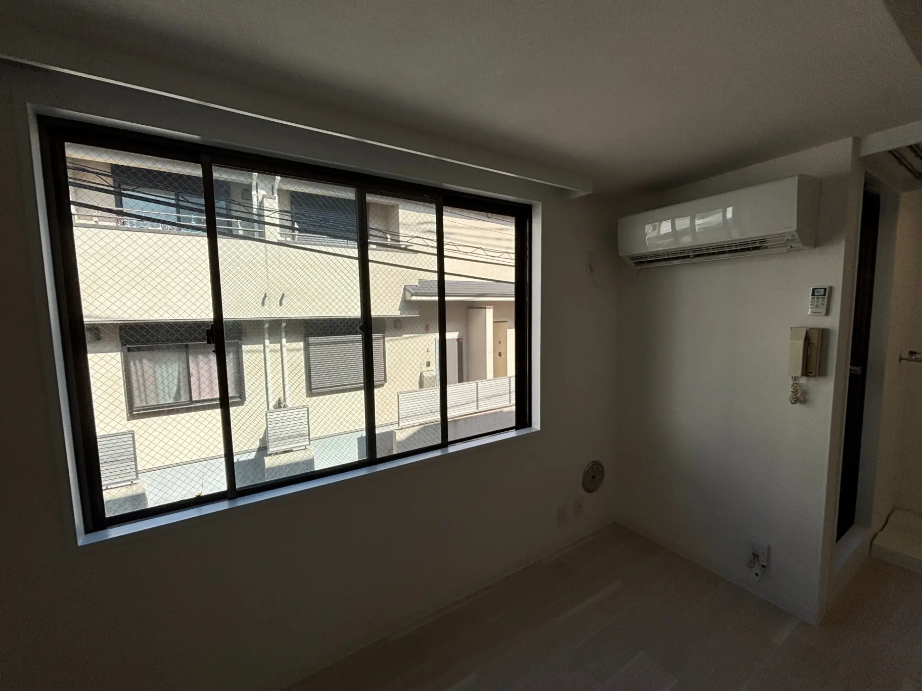 - 明るい窓から外の景色が見えるシンプルな室内、エアコンが設置された白い壁の部屋