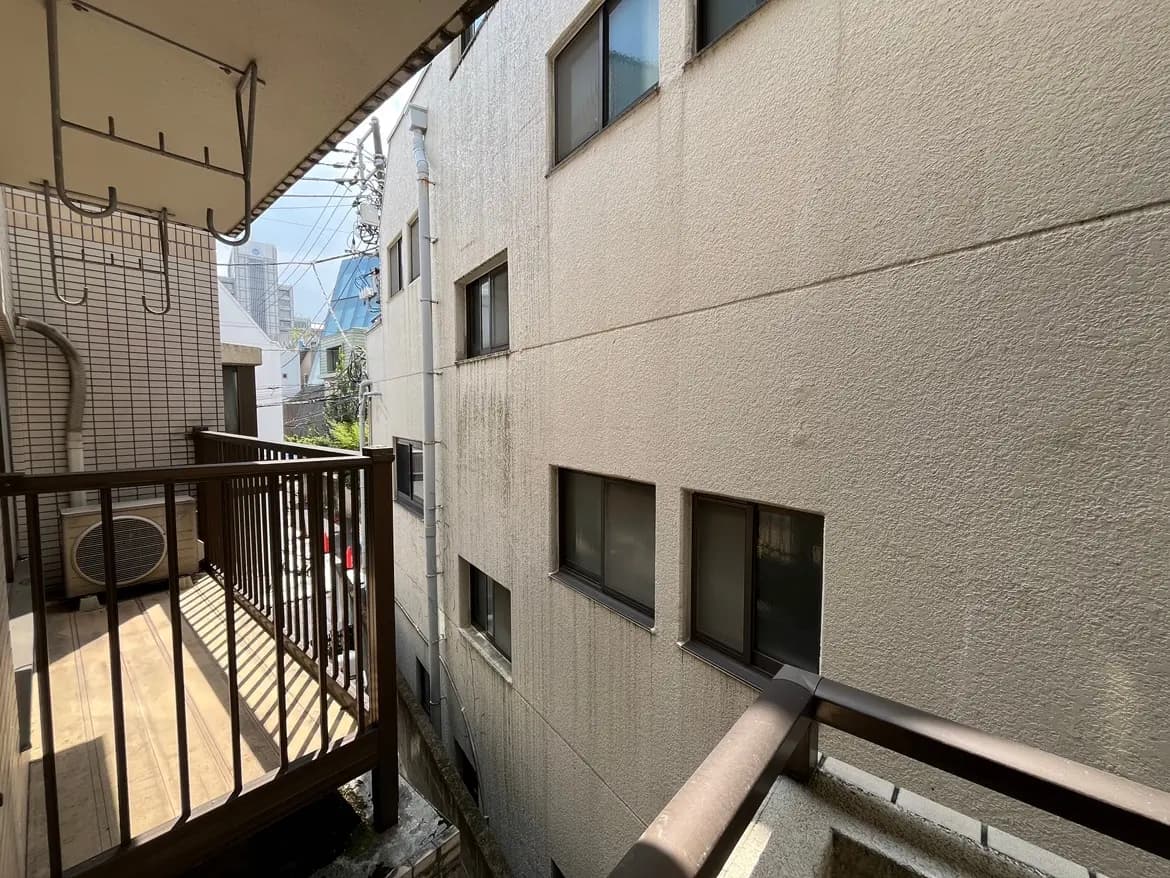 - バルコニーから見た隣接する建物の景色、手前には木製の手すりと日差しが差し込む床、ごみのない清潔感ある空間