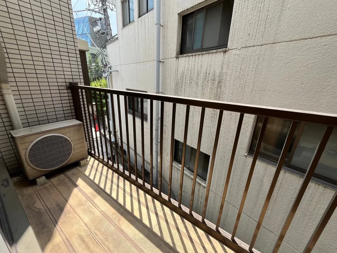 - 狭いバルコニー、木製の床にエアコン、隣接する建物の外壁が映る光景