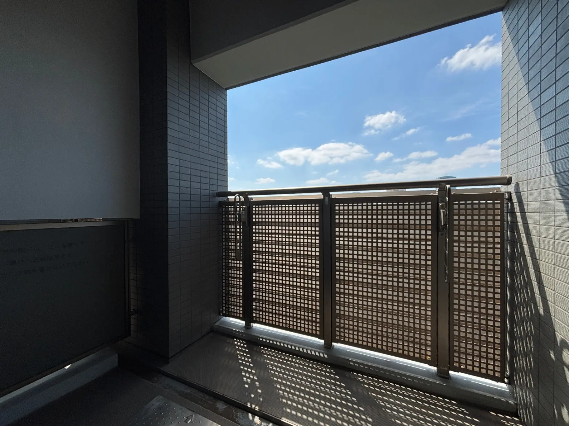 - 自然光が差し込むバルコニーの眺め、青空と白い雲が広がる景色、近くの建物が見えるシンプルなデザインの手すり