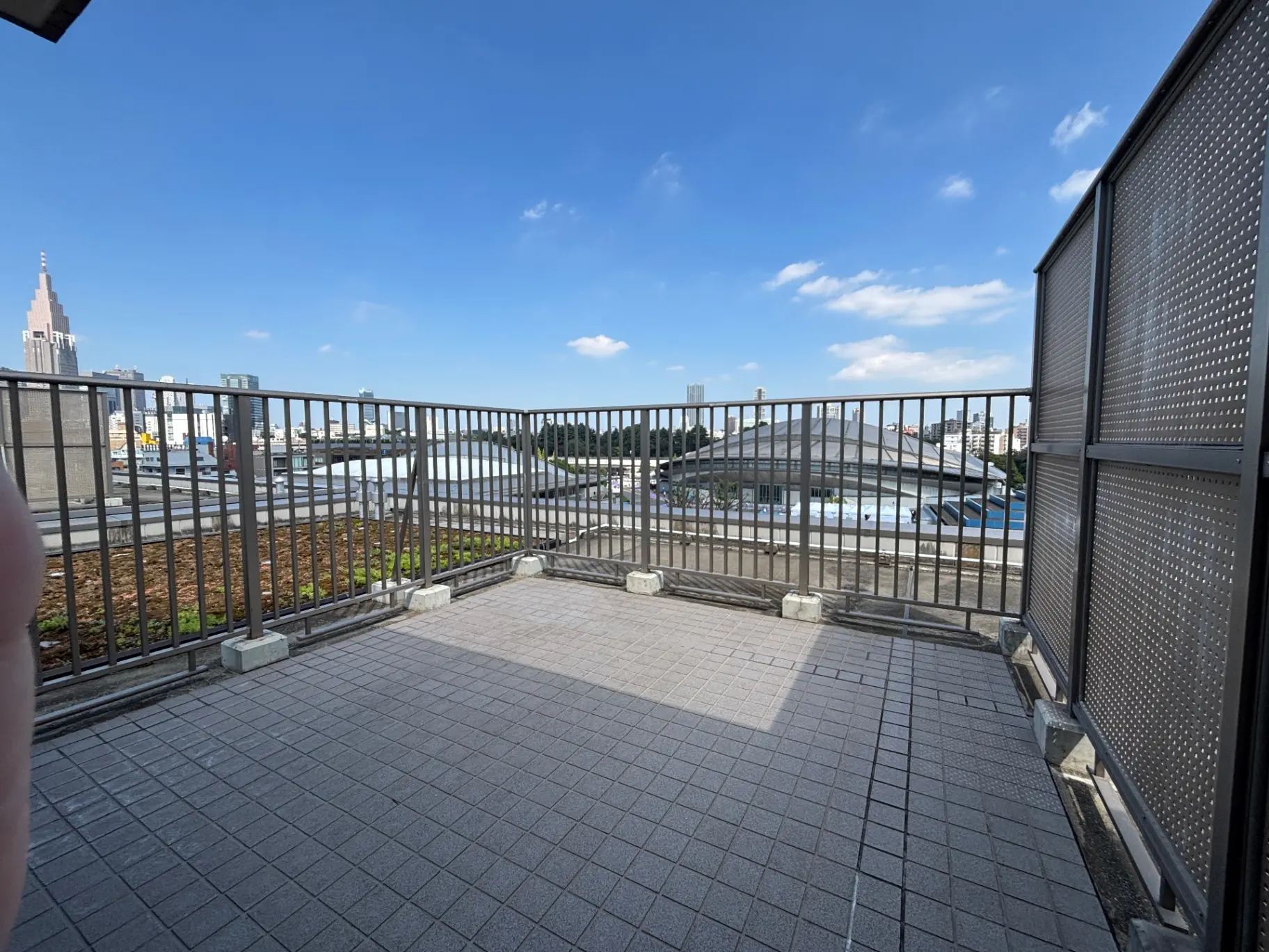 - 都市の景色を望む広々としたバルコニー、青空と雲が広がる背景、鉄製の手すりとタイルの床が特徴的な屋外スペース