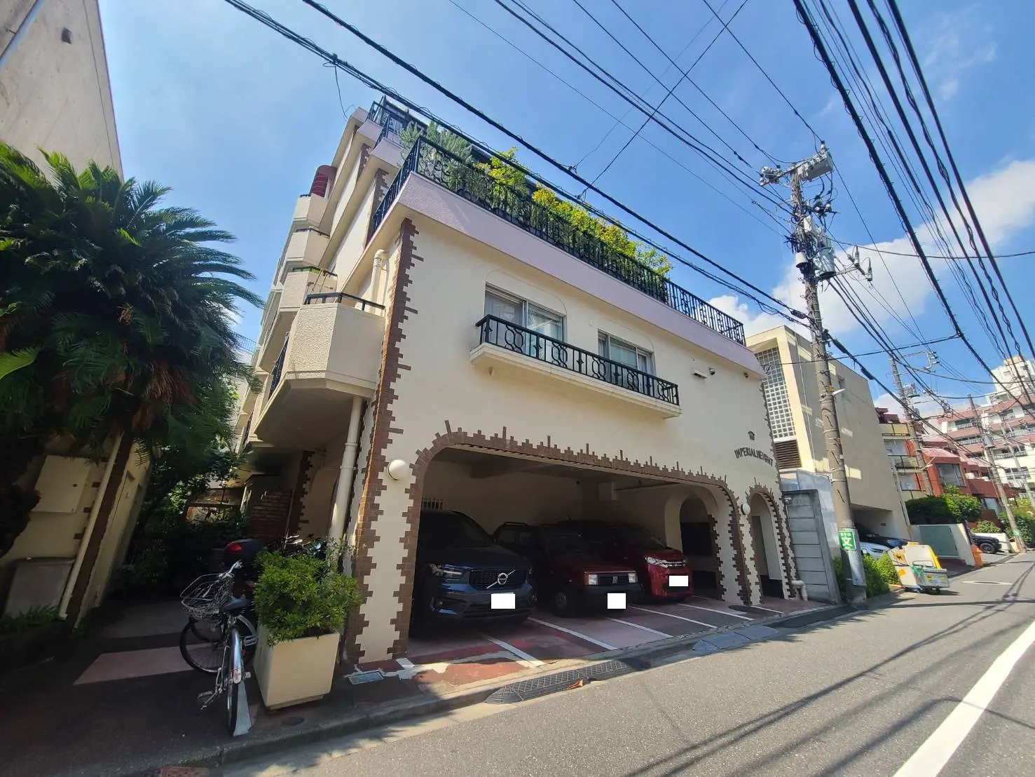 - 通り沿いの住宅、バルコニーを持つ3階建ての建物と駐車場、周囲に緑と電線が見える都市風景